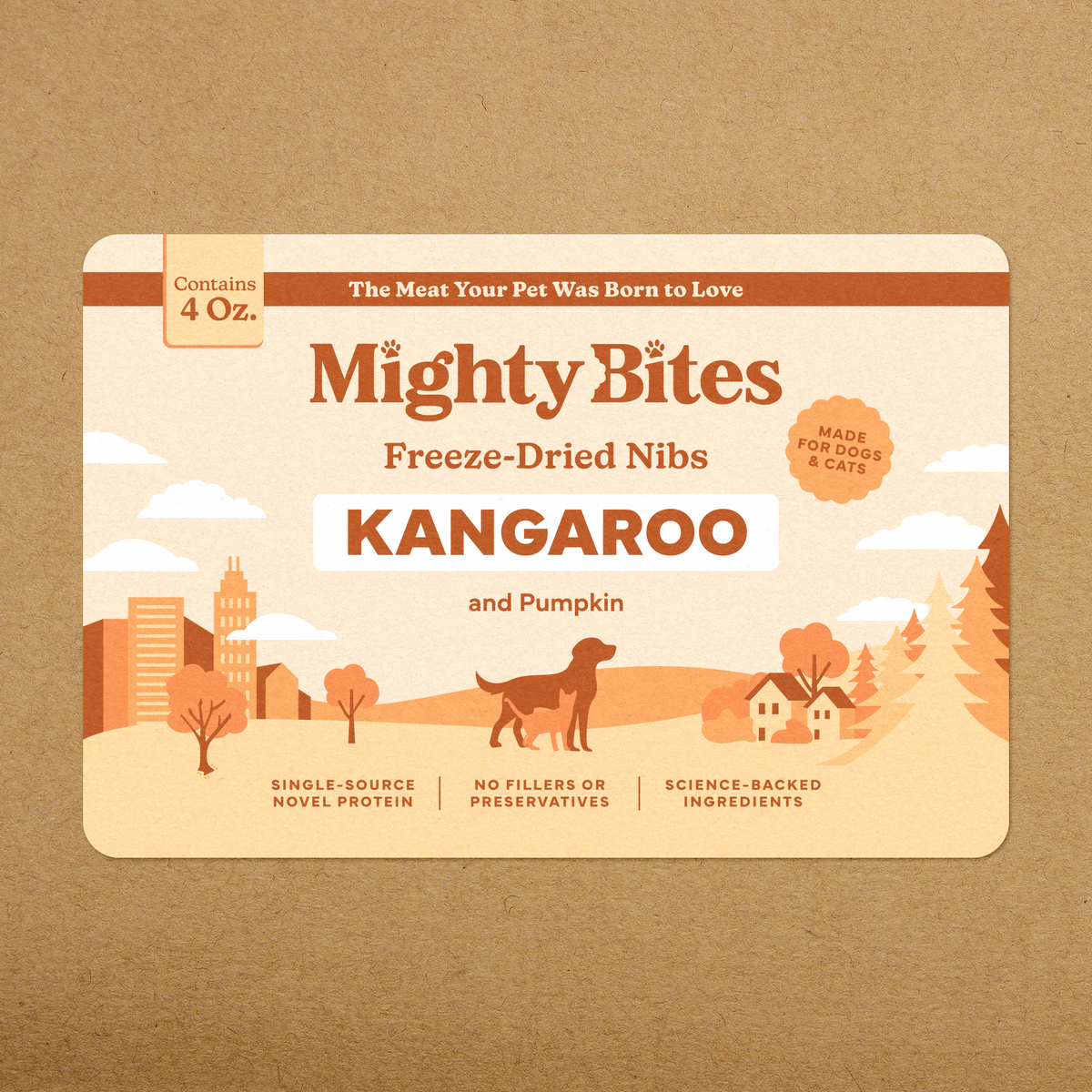 A rectangular pet food package labeled Mighty Bites Freeze-Dried Nibs Kangaroo and Pumpkin, shown flat against a brown kraft-paper background. The package is cream and orange-brown with illustrated city, trees, houses, and a dog silhouette.