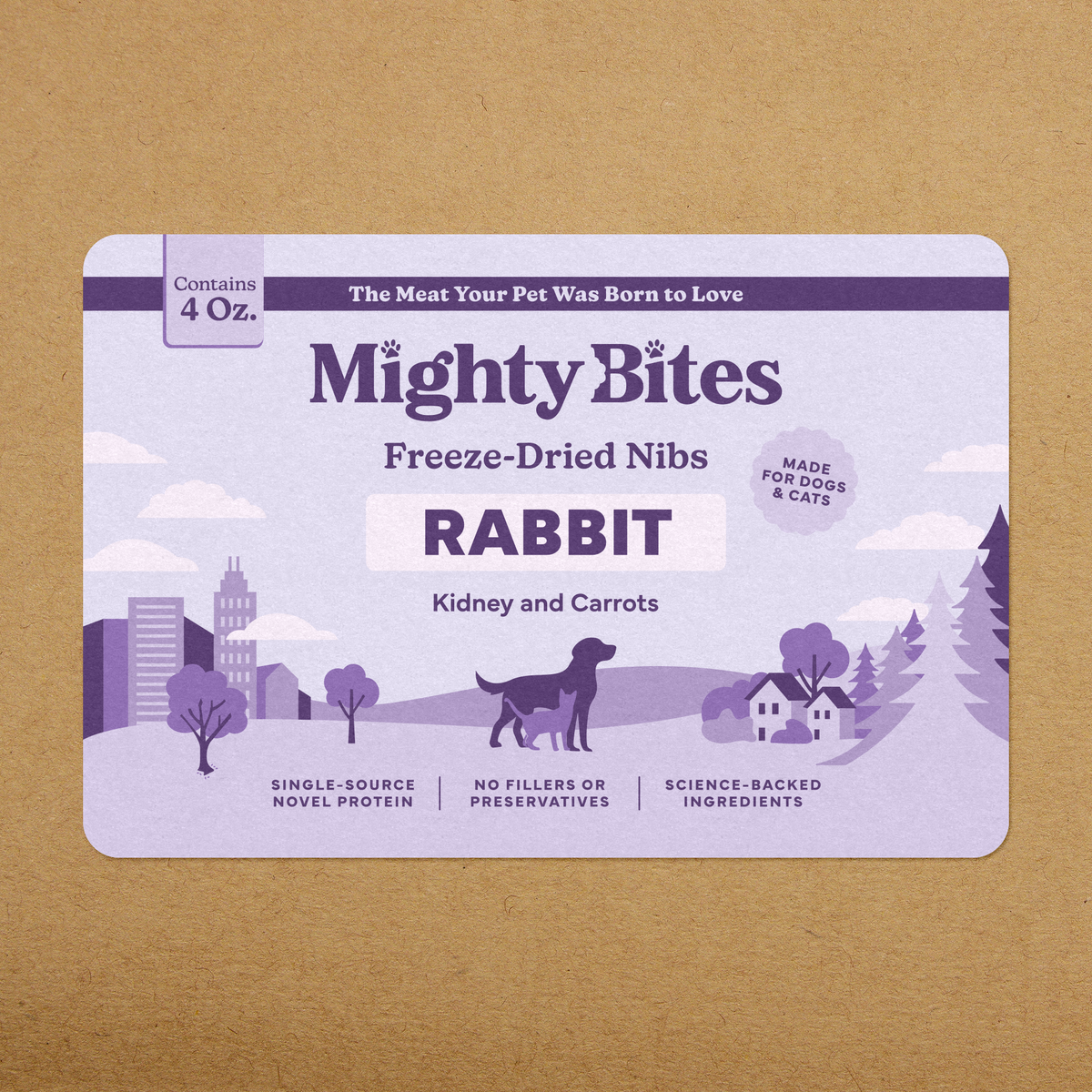 A rectangular Mighty Bites pet snack package for rabbit, kidney and carrots freeze-dried nibs, shown on a kraft paper background. The package is light lavender with purple text and illustrations, including a dog silhouette and stylized landscape.