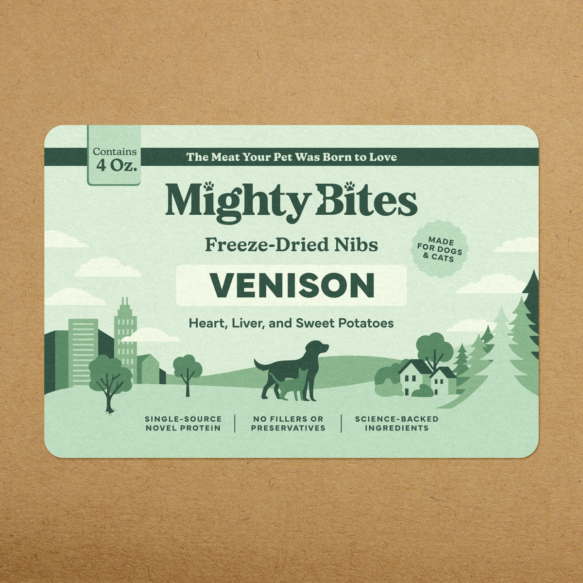 A rectangular pet food package labeled Mighty Bites Freeze-Dried Nibs Venison, shown flat against a kraft paper background. The package is pale mint green with dark green text and illustrated landscape elements, including a dog silhouette and trees.